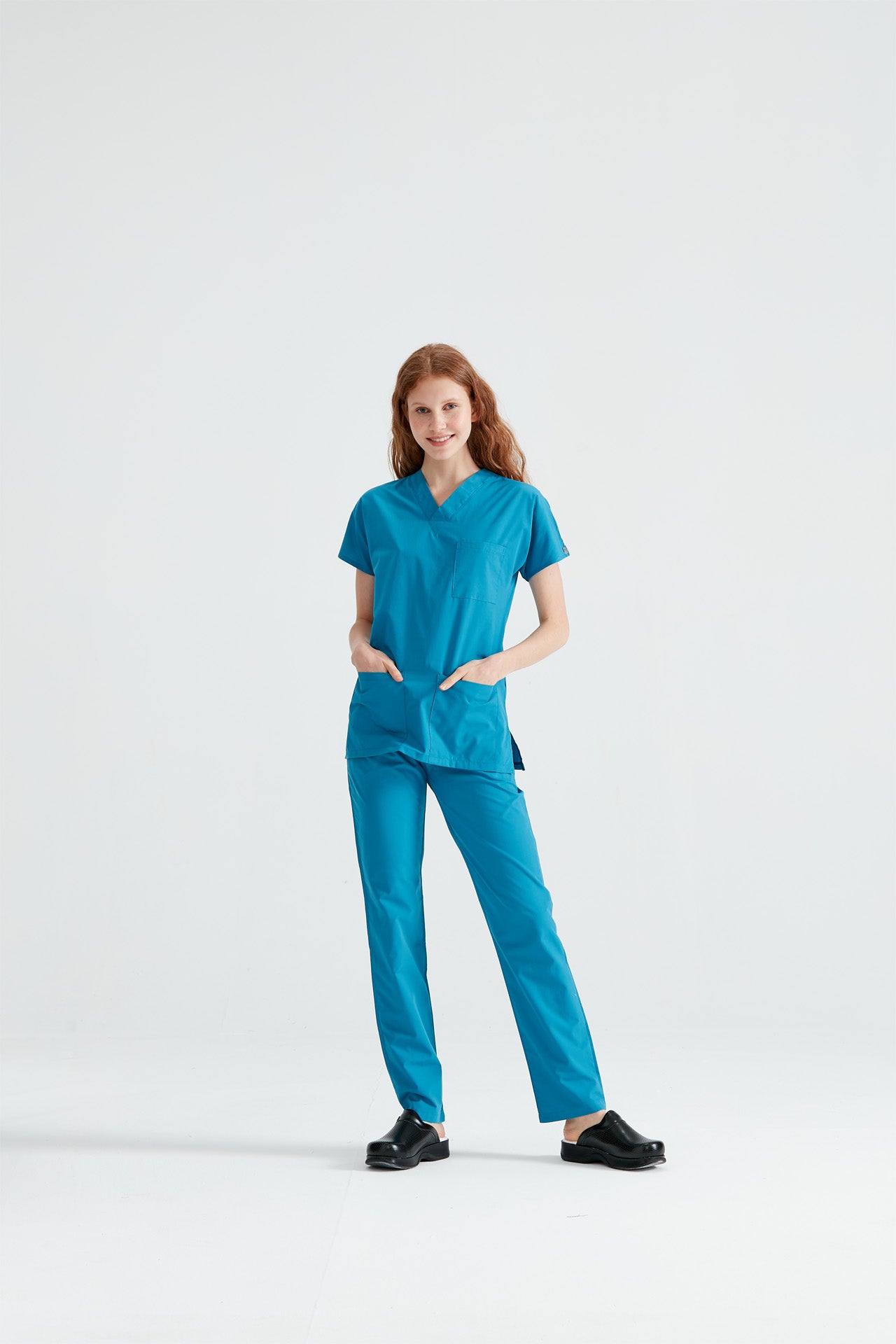 Asistenta medicala imbracata in costum medical elastan Dark Turquoise, cu mainile in buzunar, vedere din fata